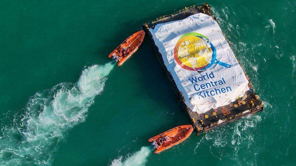 Los botes naranjas se acercan a la barcaza de socorro. Hay una hoja encima de la ayuda que dice "Cocina Central Mundial".