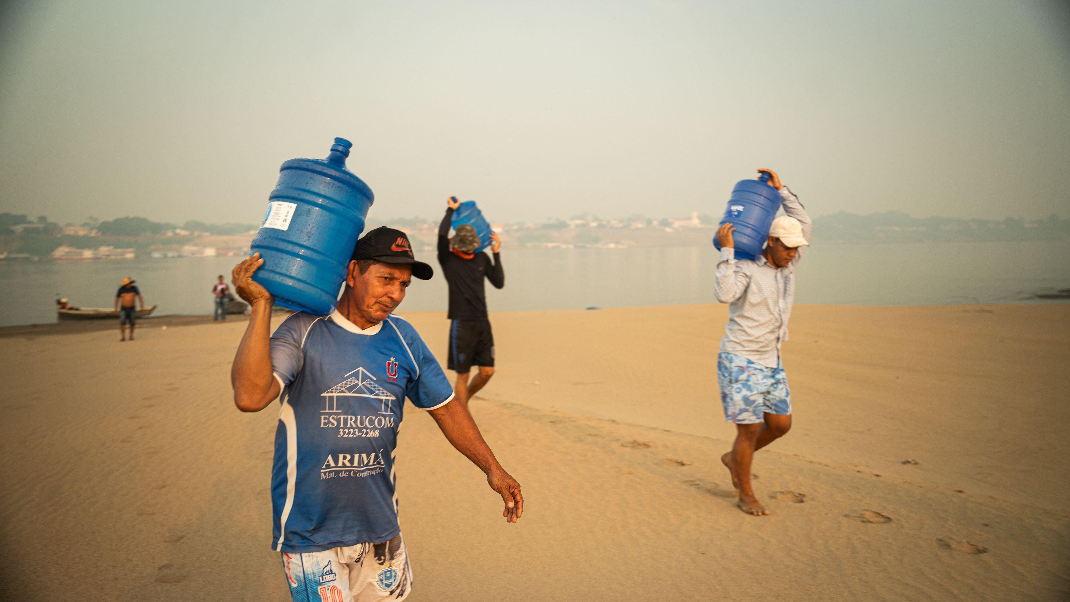 Três homens carregando galões e andando na areia