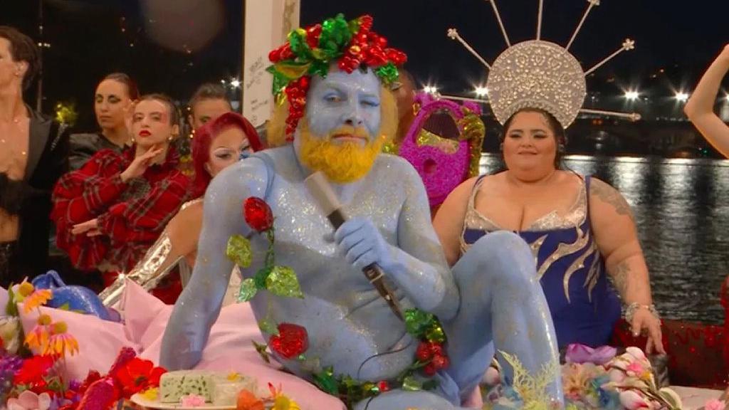 El cantante semidesnudo, pintado de azul y con una guirnalda, en una mesa y rodeado de gente disfrazada