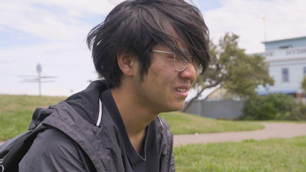 Zhang Junjie, con una camiseta y una cazadora negras, está sentado en un campo de hierba y llora. Tiene el pelo más largo que en la primera foto y lleva gafas.