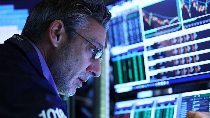 Homem olha atento para telas de computador, com gráficos e números sobre as oscilações das bolsas de valores.