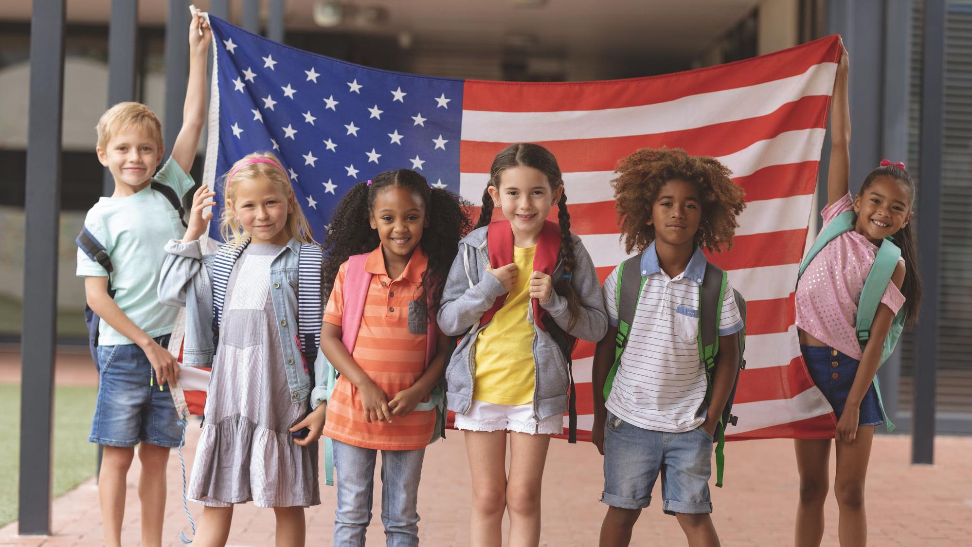 Niños estadounidenses de diversas razas y géneros frente a una bandera de EE.UU.