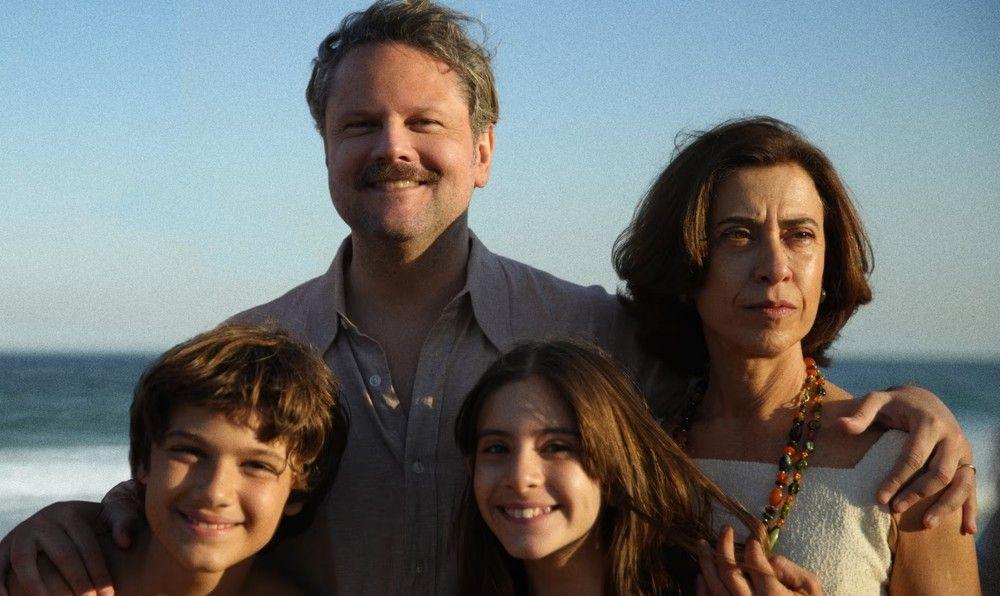 Fotografia colorida com cena do filme mostra família branca na praia: pai, mãe e duas crianças 