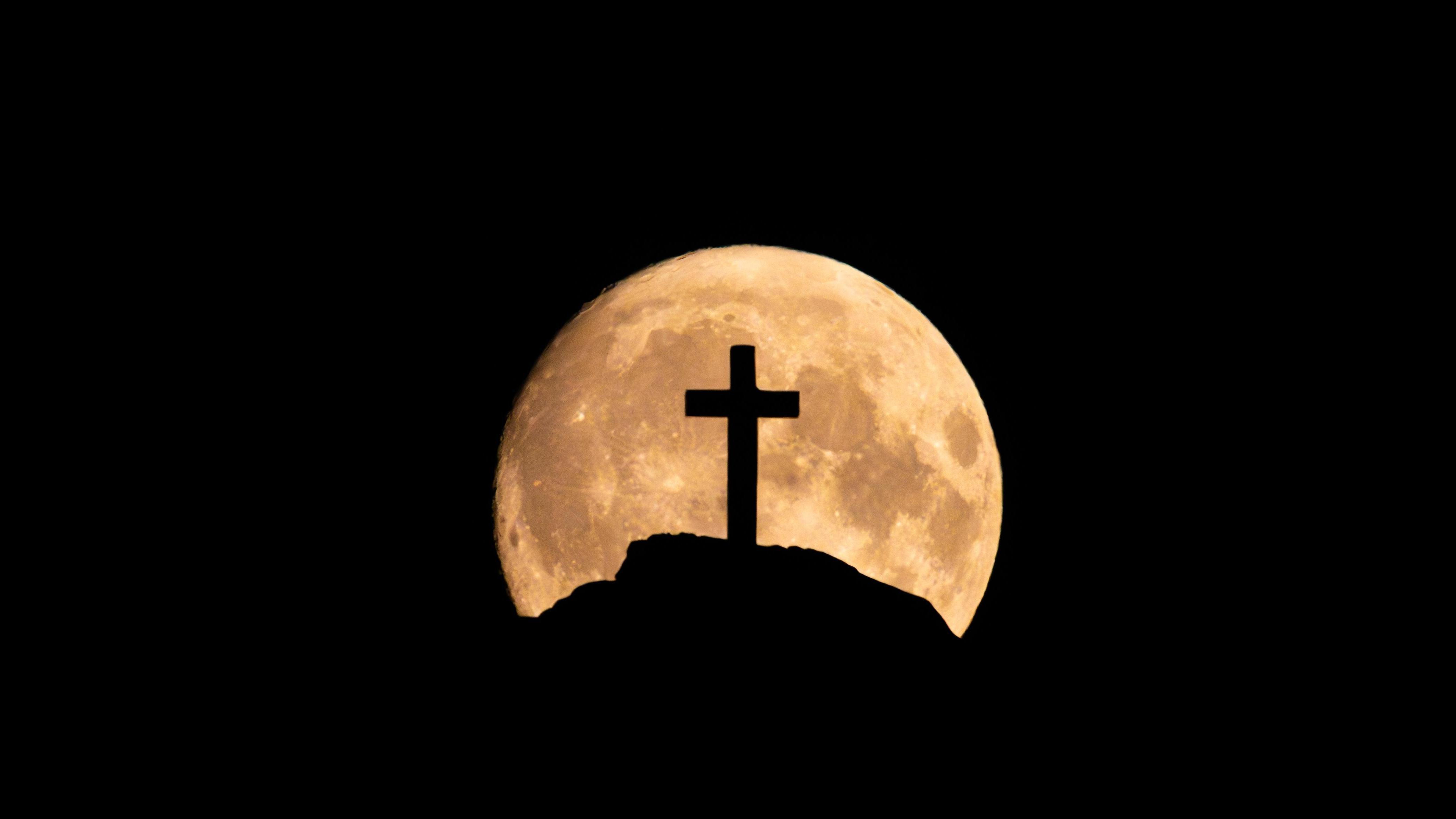 Céu escuro com a silhueta de uma colina e uma cruz religiosa no topo. Atrás da colina, a lua cheia e brilhante se destaca de forma marcante enquanto sobe no horizonte.