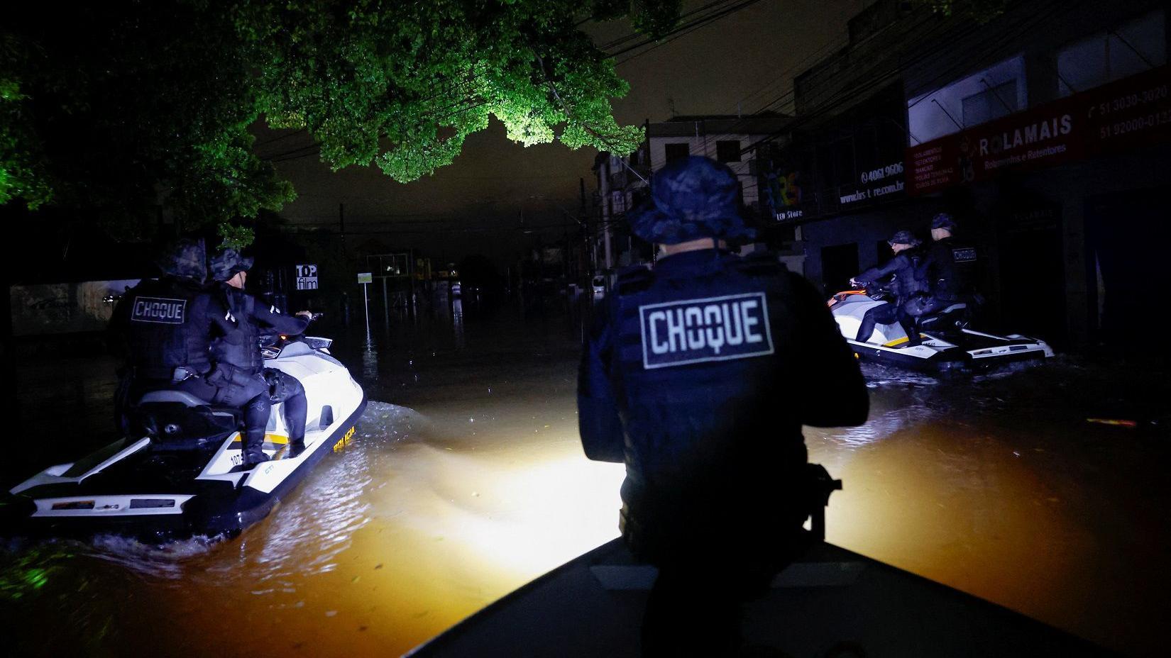 Fotografia colorida mostra policiais fazendo ronda de jet ski à noite  em uma rua alagada