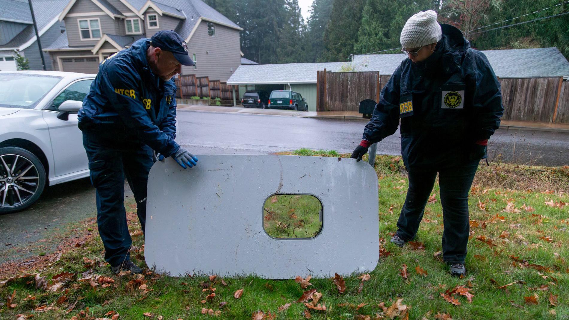Duas pessoas vestindo casacos seguram parte recuperada da fuselagem do Boeing 737 da Alaska Airlines em jardim de casa no Oregon