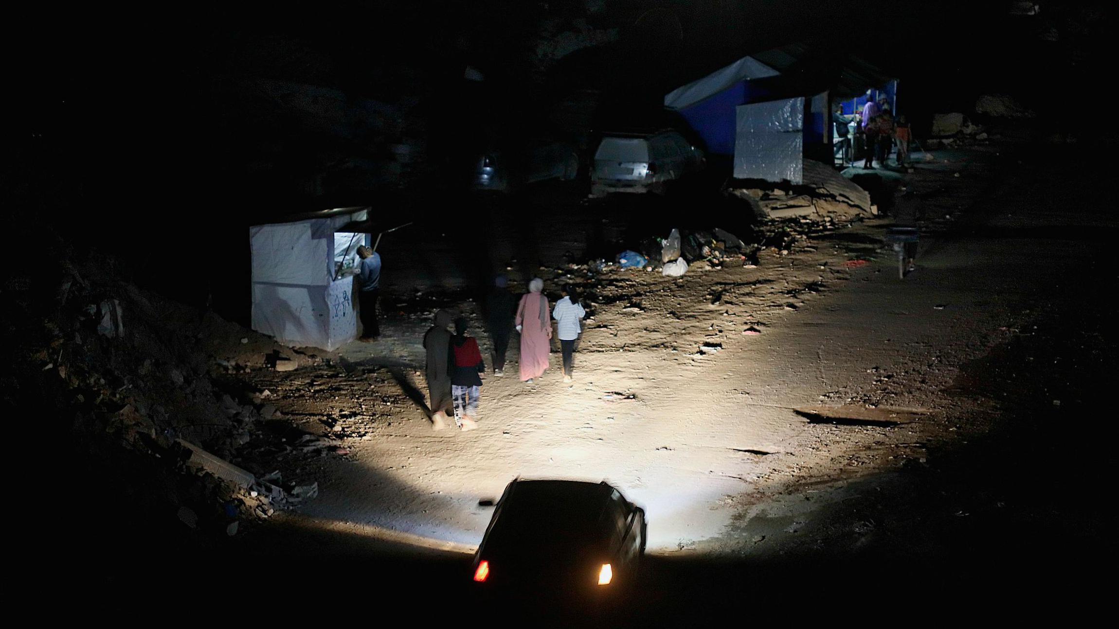 Mujeres caminan por una calle oscura iluminada por los faros de un vehículo durante los cortes de electricidad en la Franja de Gaza.