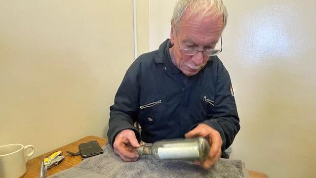 Barry examina a garrafa. Ele está sentado à mesa, tem cabelos brancos, usa óculos e um uniforme de trabalho azul