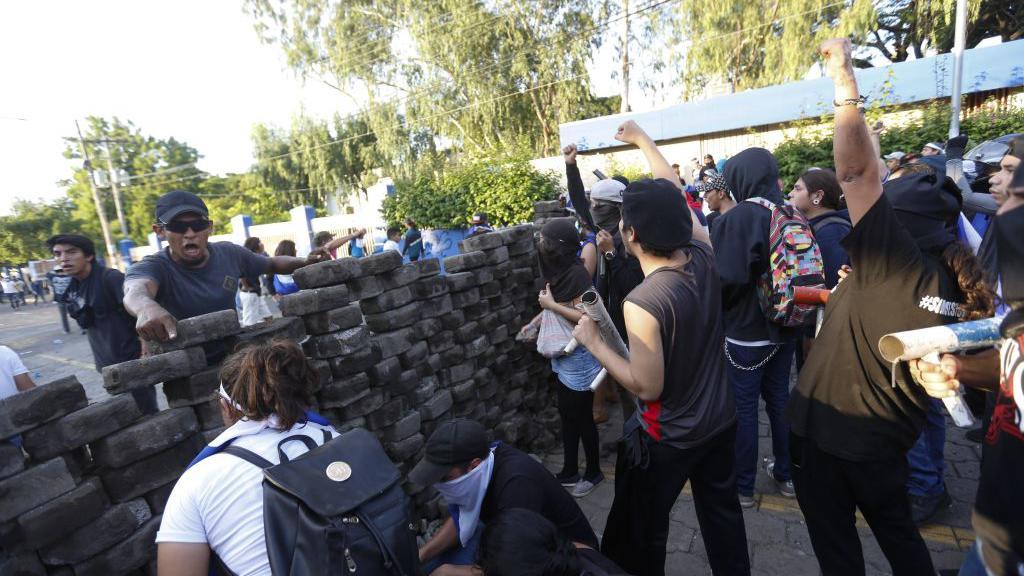 Pessoas erguendo os braços protestando atrás de barricada; do outro lado, aparece homem gritando e apontando para manifestantes