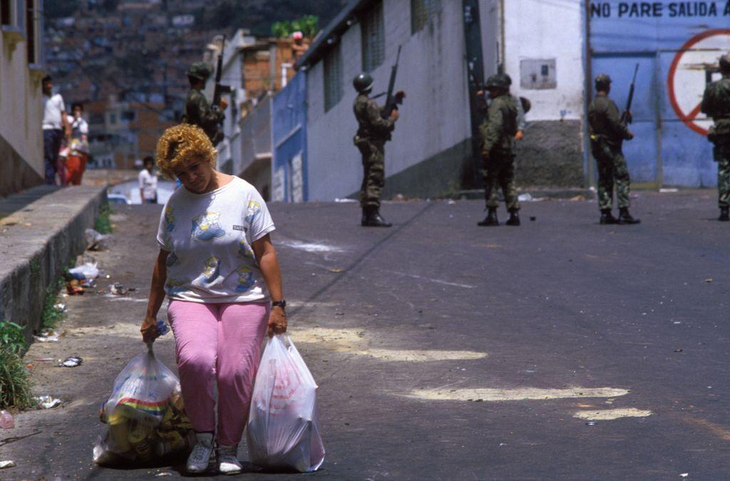 Una mujer camina cargada con bolsas, con tres militares al fondo en una calle de Caracas, en una imagen de 1989.