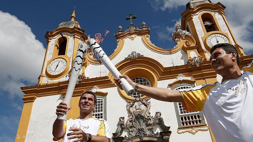Dos atletas hacen relevo de la antorcha olímpica frente a una Iglesia en Brasil en 2016