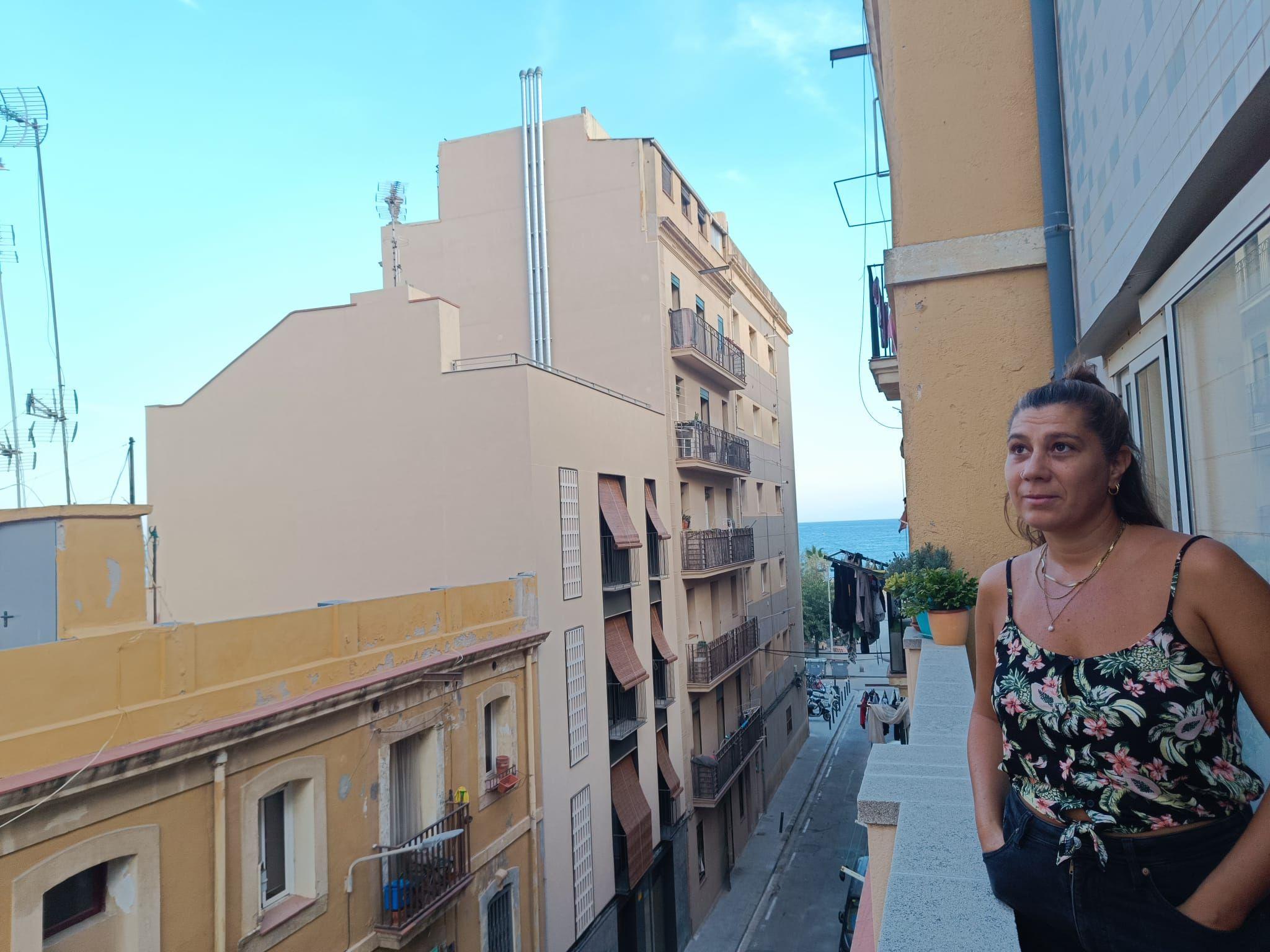 Maíra olhando para o lado da varanda de seu apartamento, com rua abaixo e vista para o mar ao fundo