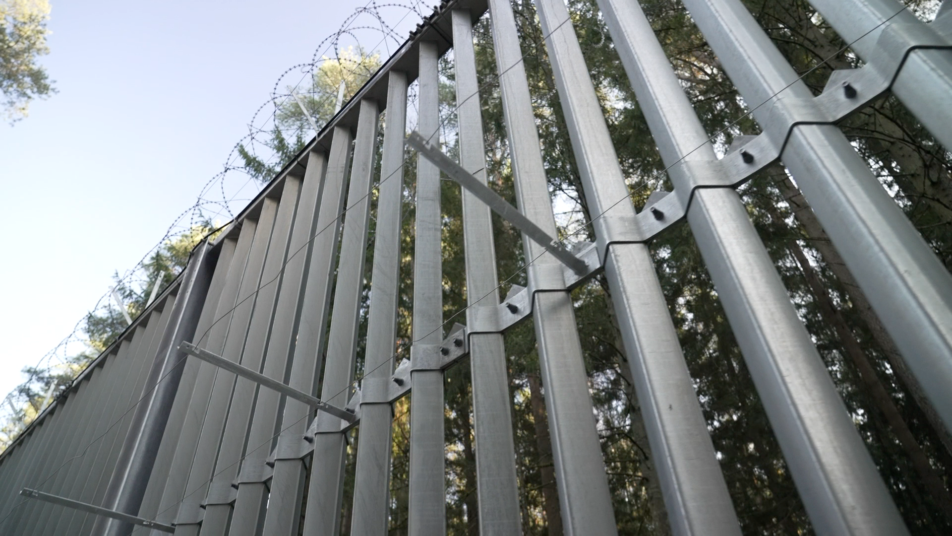 A tall silver palisade fence in the sunshine, topped with rolls of barbed wire.