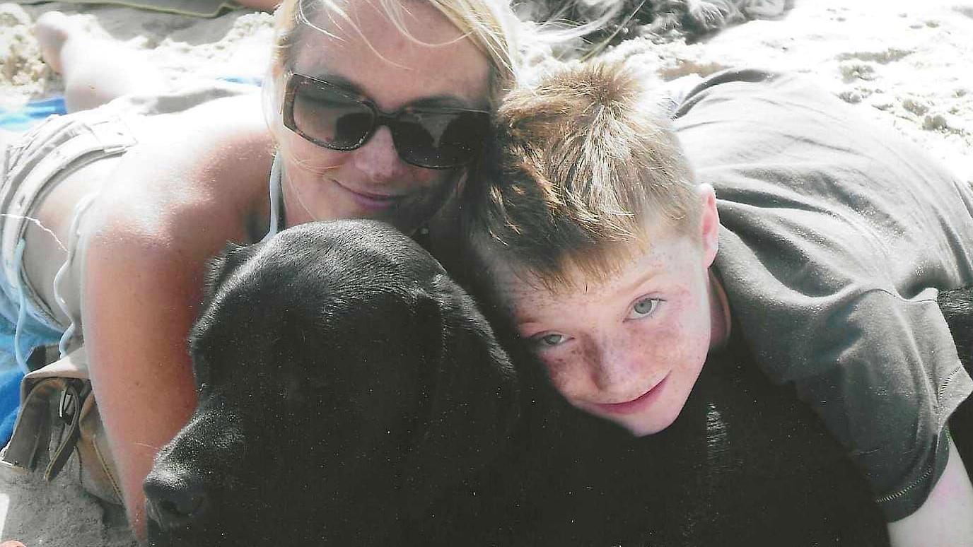 Declan, ainda criança, deitado na praia com sua mãe, Alex, e seu cachorro