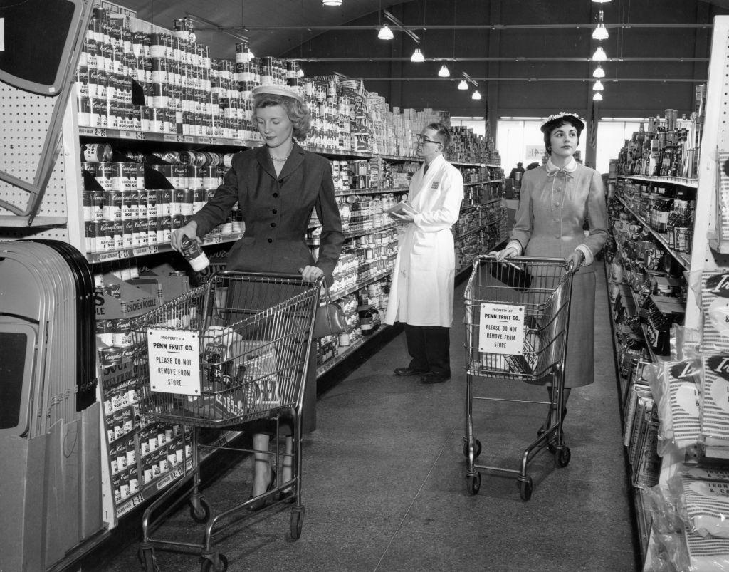 Dos mujeres de compras en un supermercado en Estados Unidos mientras un empleado revisa los anaqueles, en torno al año 1955.