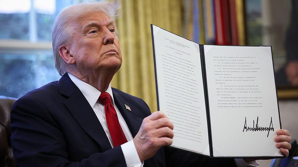 O presidente dos EUA, Donald Trump, com a boca fechada, segura uma ordem executiva assinada no Salão Oval da Casa Branca em 26 de março de 2025 em Washington, DC. O presidente Trump anunciou tarifas de 25% sobre todos os carros fabricados no exterior