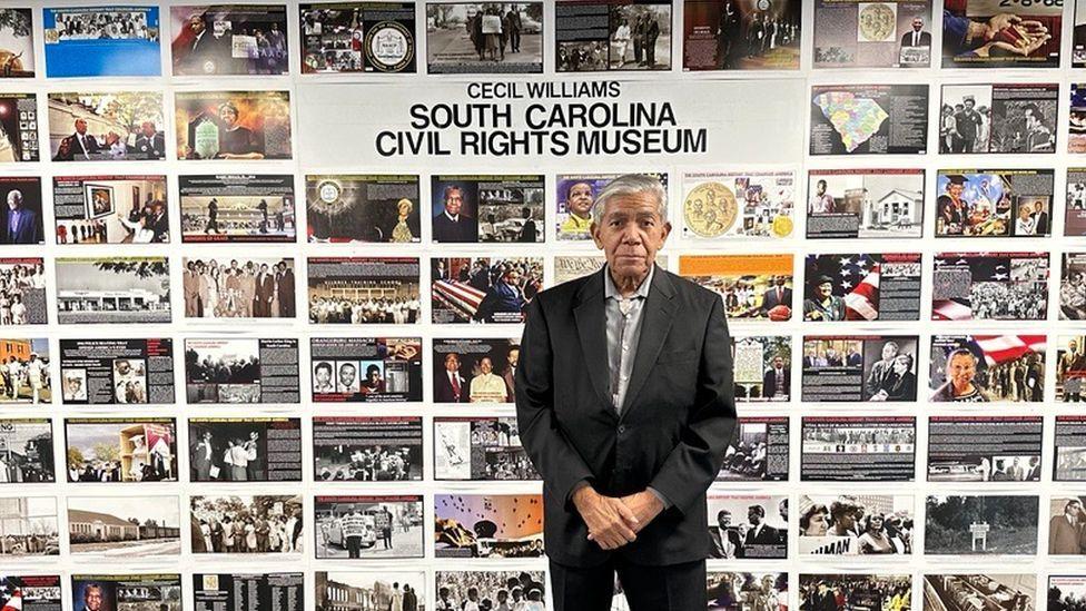 Cecil Williams em frente a parede de fotos históricas no Museu dos Direitos Civis da Carolina do Sul