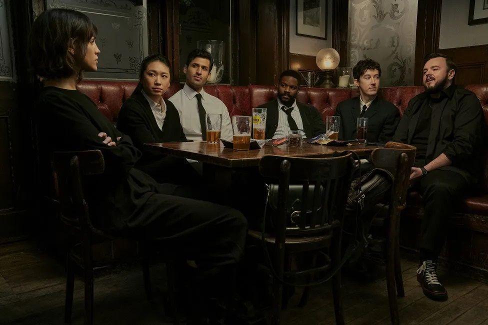 Cena da série em que mostra cinco cientistas e oficial da Marinha sentados à mesa de um bar