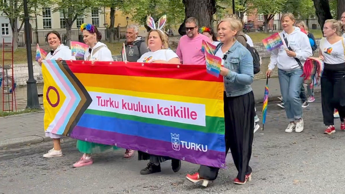 Una marcha en Turku, con un grupo de líderes de la ciudad marchando al frente y sosteniendo una bandera arcoíris. Las personas que hablan también sostienen banderas LGBTQ+ mientras caminan.