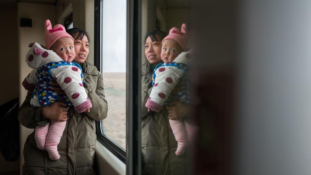 A Chinese woman holds her baby in her arms next to the window of a train. 