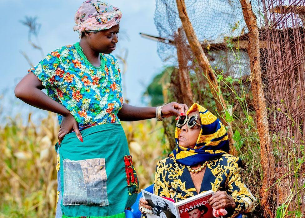 Diana Kaumba está do lado de fora, vestindo roupas tradicionais da Zâmbia, olhando para sua avó sentada, que usa uma jaqueta de grife com estampas azul-marinho e dourada, cachecol e óculos de sol. Ela segura um livro intitulado Hollywood Fashion, com Audrey Hepburn na capa.