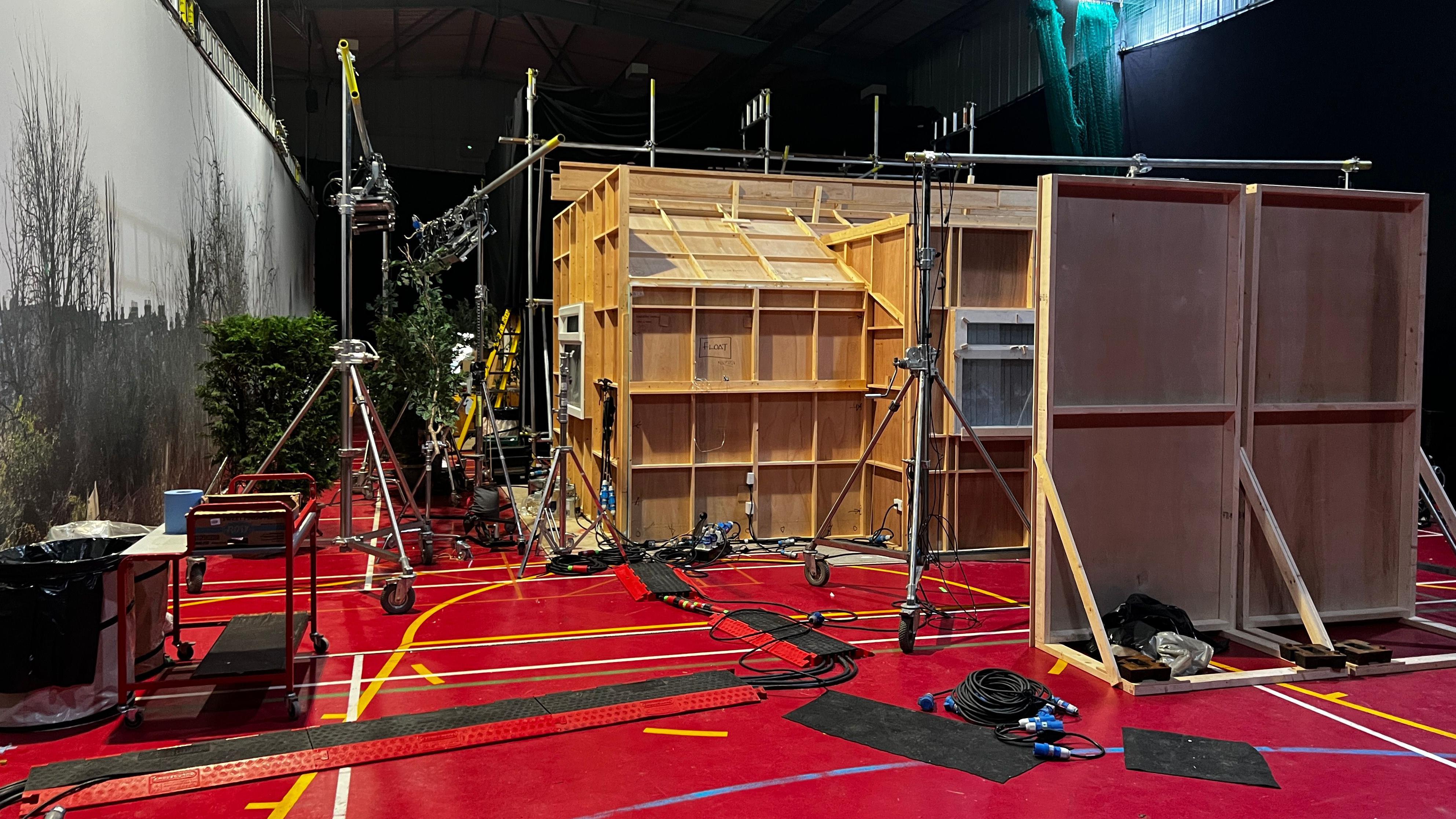 Parte externa de cenário de filme, sustentada por andaimes. É uma caixa de madeira com janelas, e do lado de fora das janelas há árvores e arbustos falsos. O chão está coberto de cabos e equipamentos de filmagem.