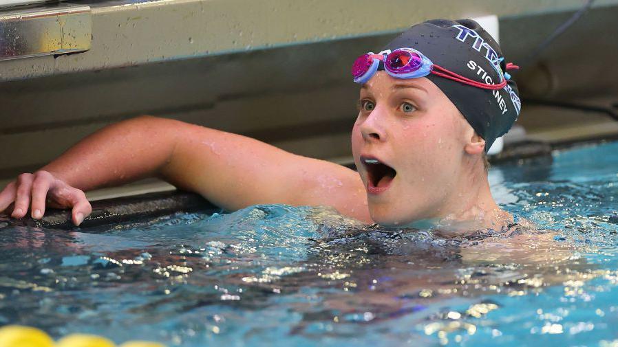 A foto mostra uma mulher de pele e olhos claros, com touca e óculos de natação, em uma piscina, celebrando.