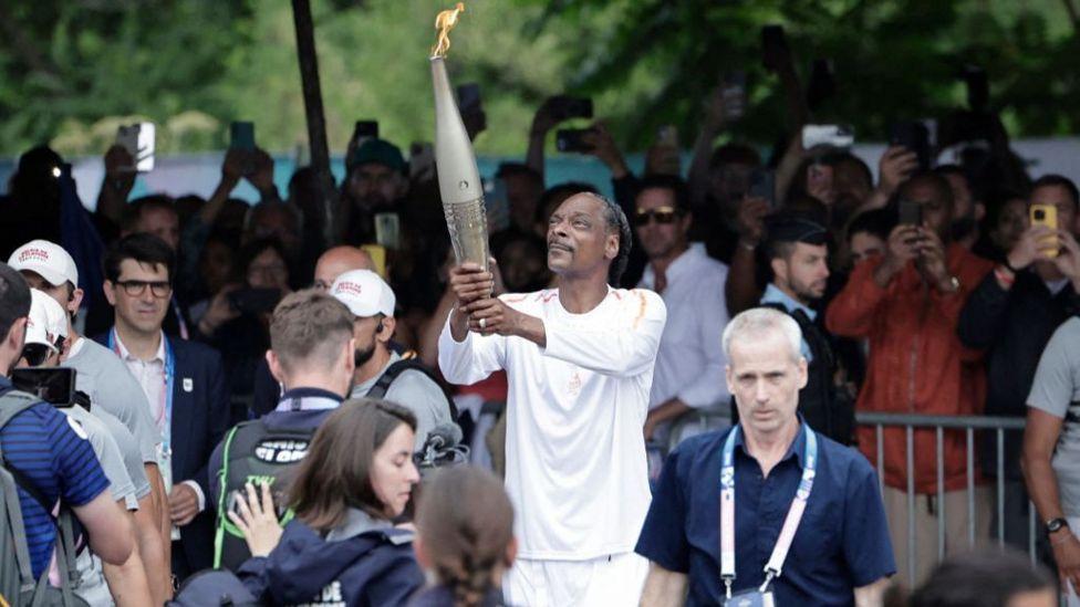Snoop Dogg vestido con ropa deportiva blanca y llevando la antorcha olímpica. 