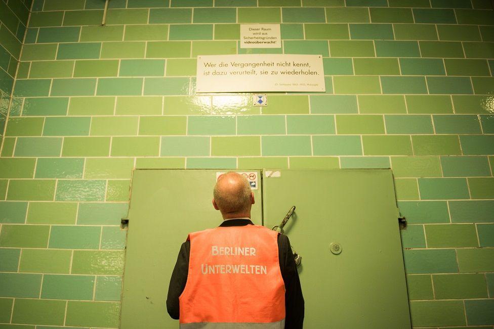 Dietmar Arnold de costas em frente a uma instalação de um bunker da Segunda Guerra Mundial. Acima, há uma placa com a frase de Santayana