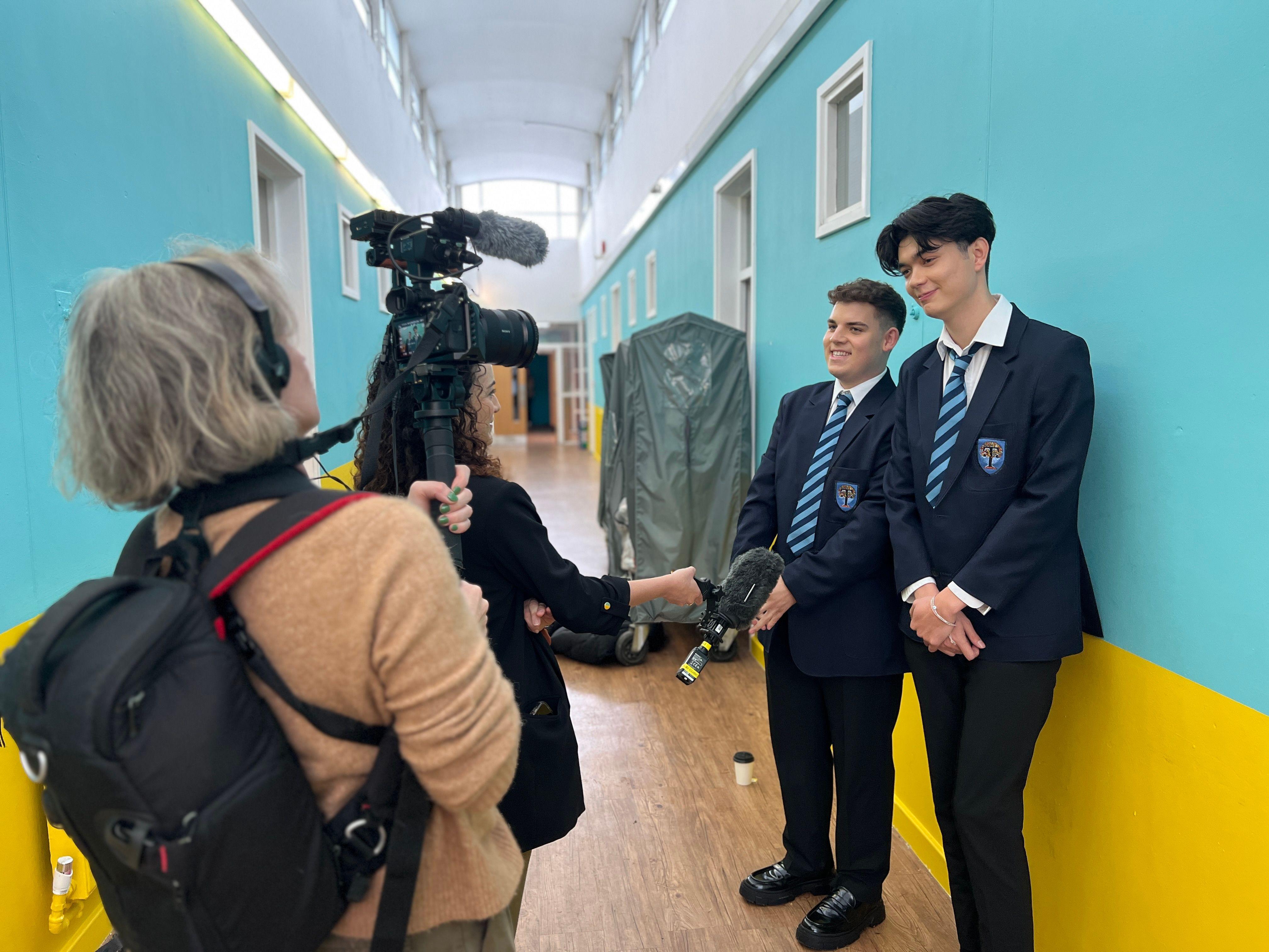 Uma operadora de câmera com uma mochila de equipamento nas costas filma Tobie Donovan e William Gao, enquanto uma repórter estende um microfone para entrevistá-los. Os dois estão rindo.