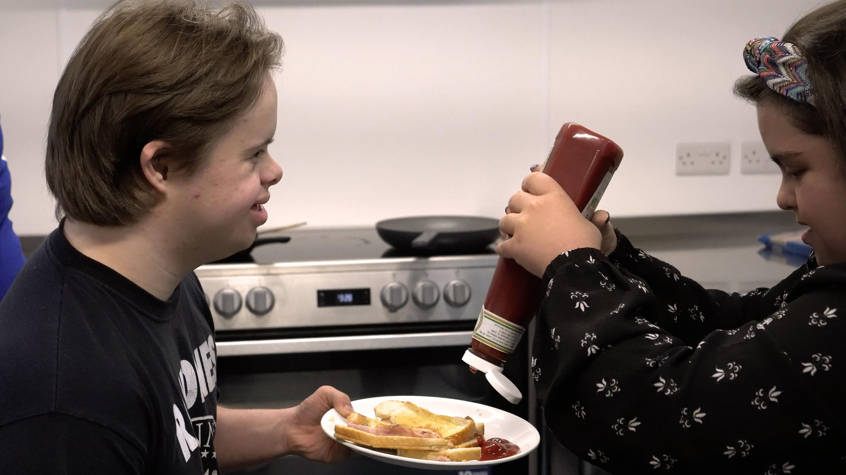 Dois jovens com síndrome de Down, um deles segurando um prato com um sanduíche e o outro colocando ketchup no prato