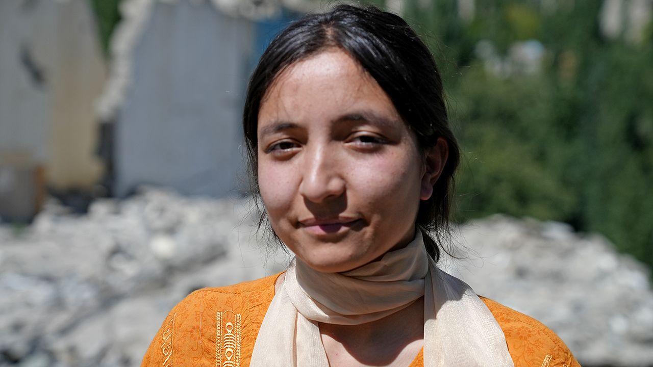 A close up head and shoulders portrait of Komal Sher wearing a white scarf and orange top, looking at the camera pictured with rubble in the background in her village of Hassanabad.