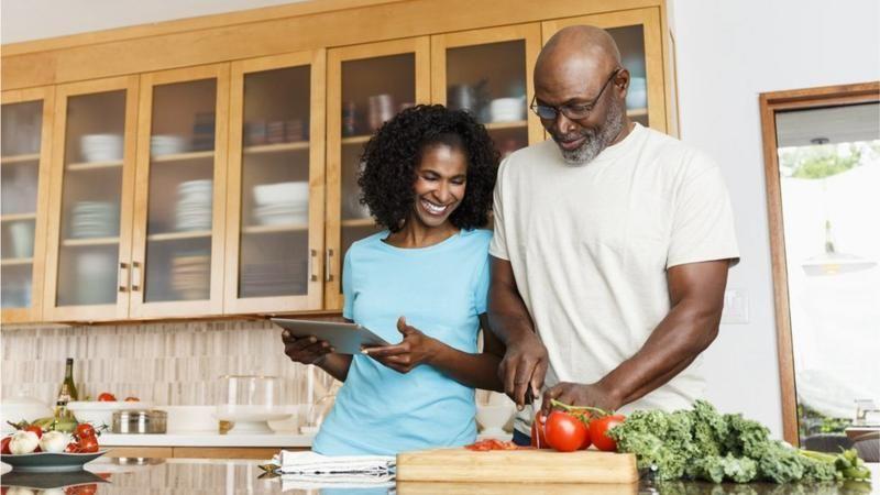 Casal cozinha juntos; a mulher segura um tablet enquanto o homem corta tomates