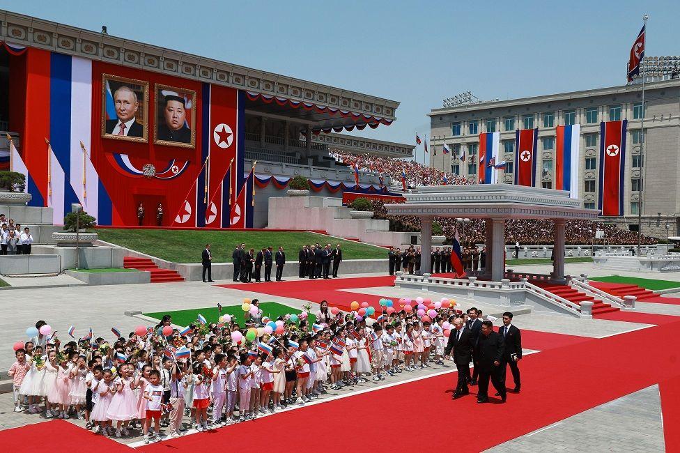 Vladimir Putin, presidente de Rusia, camina junto a su homólogo de Corea del Norte, Kim Jong-un, y pasan al lado de un grupo de niños. De fondo se ven fotos de los dos líderes