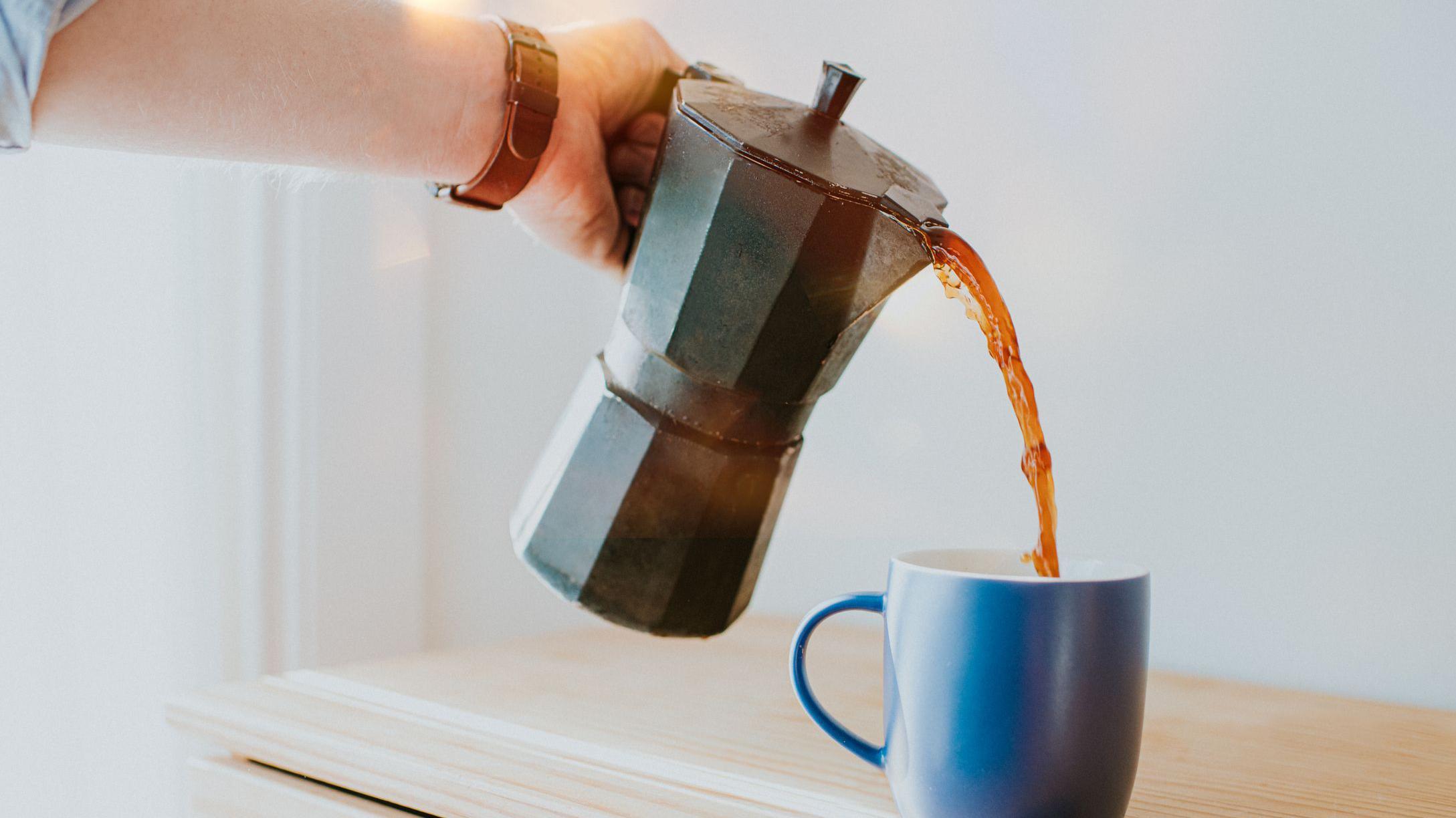 Mão masculina servindo café com uma cafeteira italiana em uma caneca azul