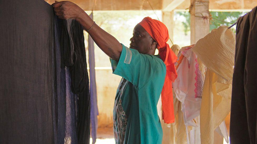 Perempuan dengan baju hijau dan kerudungmerah menggantungkan kain hitam yang telah dicelup di Mbacke Kadior