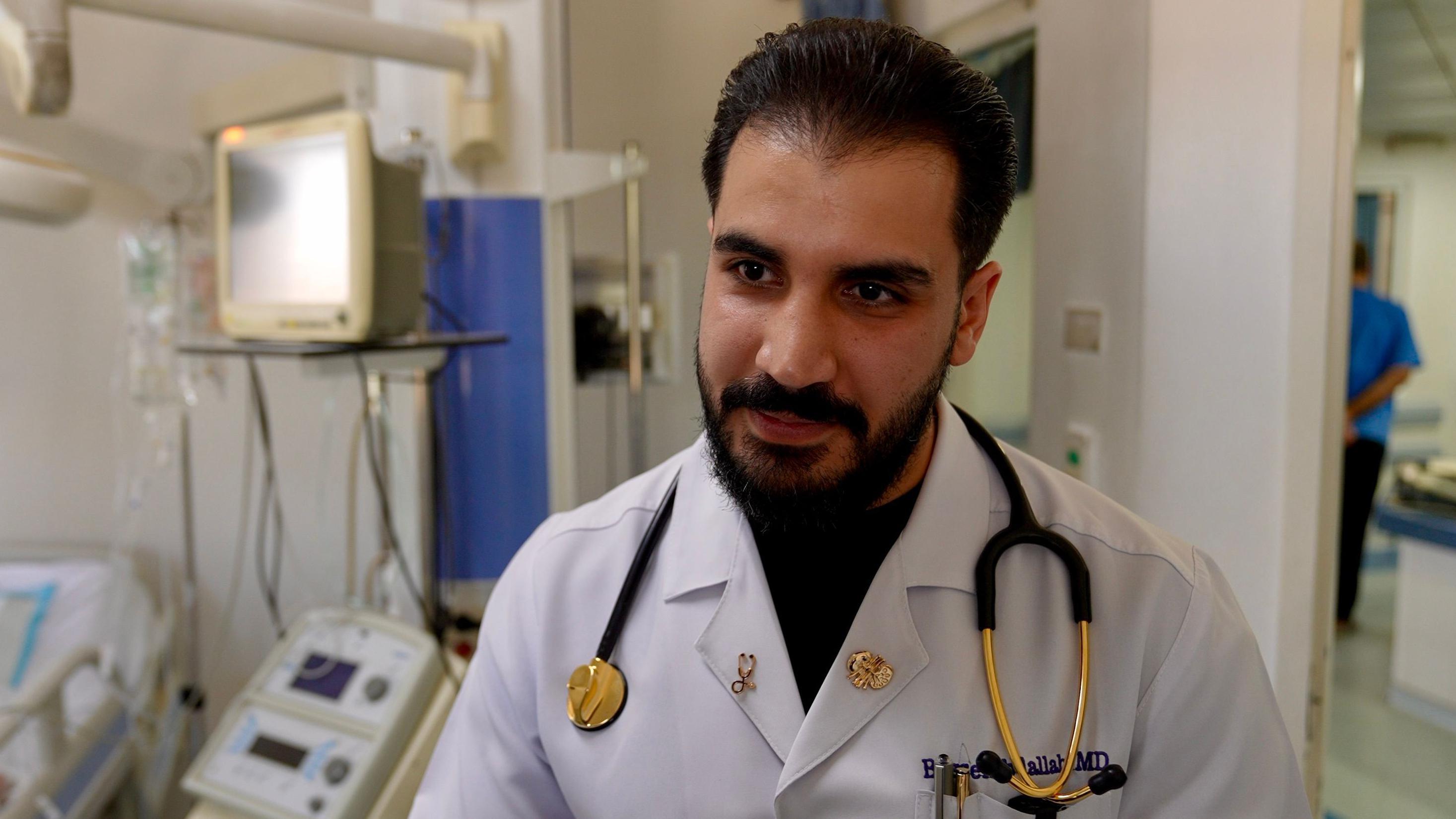 Basil Abdallah, com barba e cabelos pretos, vestindo um jaleco branco e um estetoscópio pendurado no pescoço, no hospital.