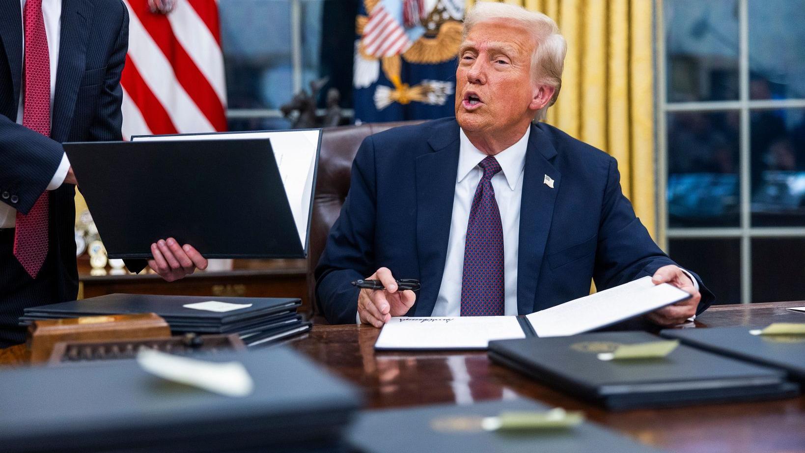 Trump sentado no Salão Oval com uma mesa cheia de pastas com os decretos que assinou no primeiro dia de seu segundo mandato