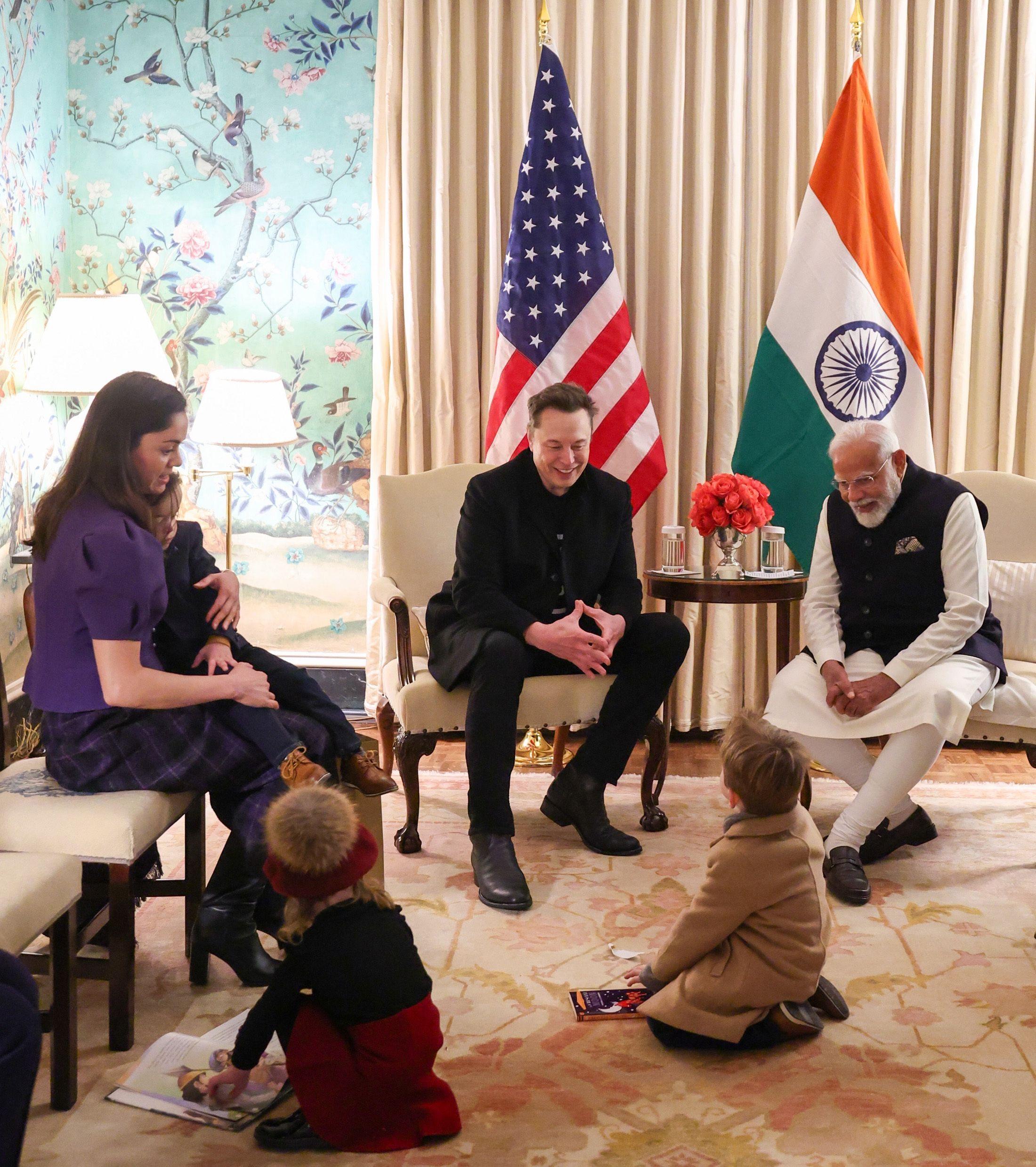 Elon Musk sentado ao lado do primeiro-ministro indiano, Narendra Modi, com duas crianças brincando no chão. Há bandeiras dos EUA e da Índia visíveis atrás deles