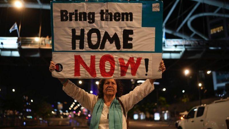 Mulher com cartaz escrito 'Traga-os para casa agora', em inglês