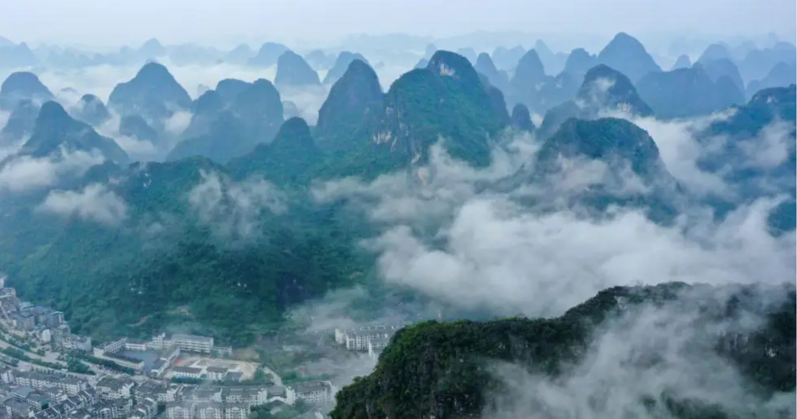 A imagem mostra uma paisagem natural composta por montanhas cobertas de vegetação, com picos pontiagudos que emergem de uma espessa neblina, criando uma atmosfera etérea. Ao fundo, encontra-se um pequeno aglomerado urbano com edifícios e ruas, que contrasta com a imensidão das formações naturais. O céu apresenta tons azulados, reforçando a atmosfera de serenidade e mistério do cenário.