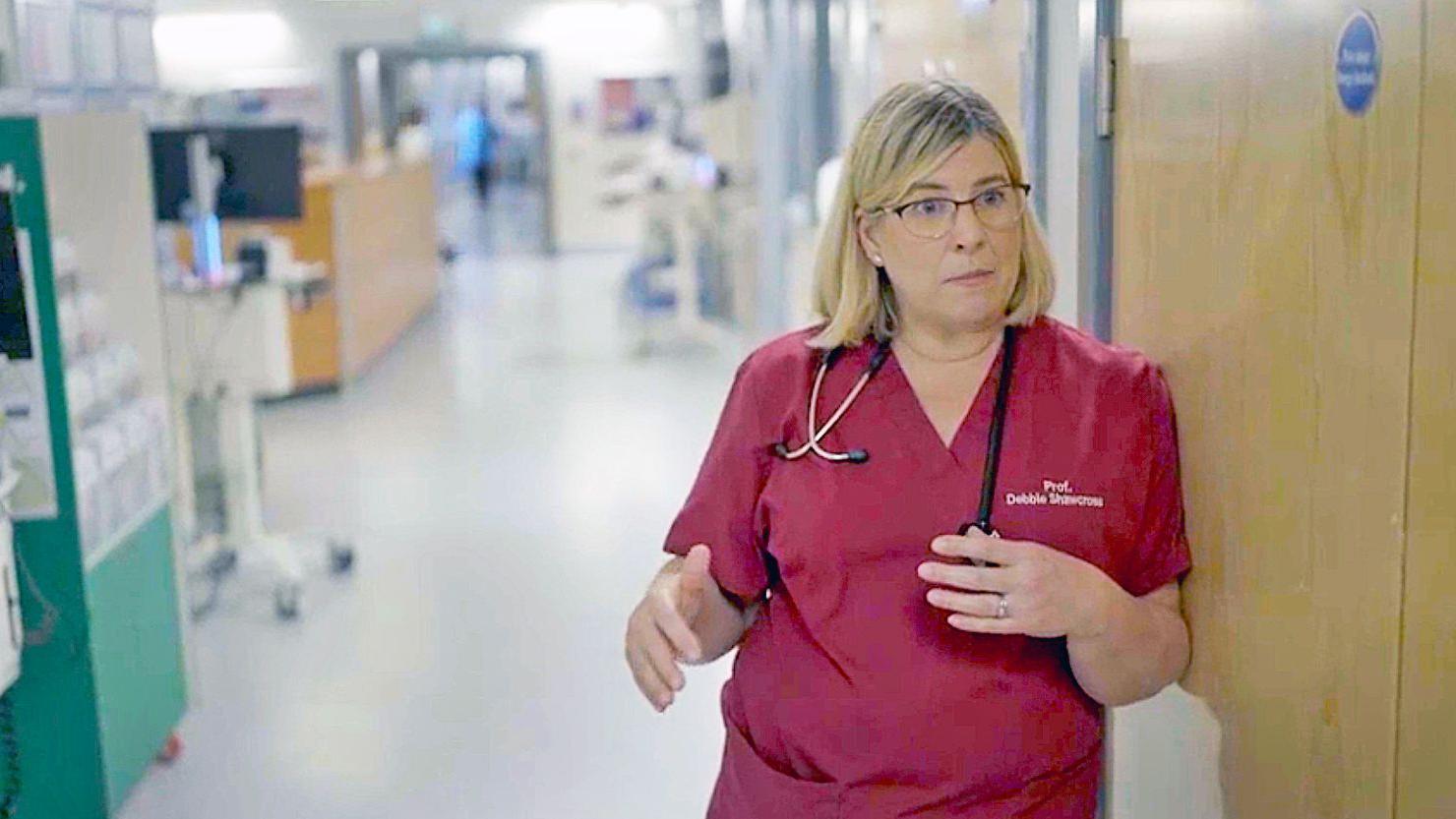 A hepatologista consultora Debbie Shawcross, vestindo um uniforme vermelho e um estetoscópio pendurado no pescoço, em pé no corredor de um hospital.