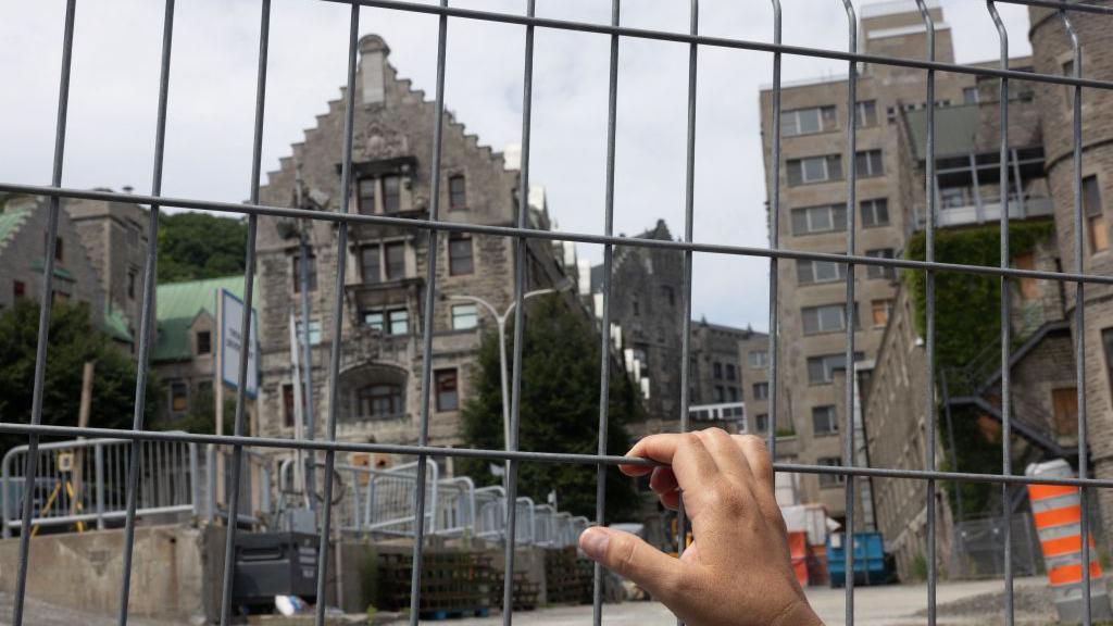 A mãe Mohawk, Kwetiio, olha através de uma cerca no canteiro de obras do Royal Victoria Hospital em 17 de julho de 2024, em Montreal, Canadá. Uma mão segura uma cerca de metal em frente a um antigo prédio de pedra, em uma área em construção com máquinas, grades e cones laranja, sob um céu nublado.