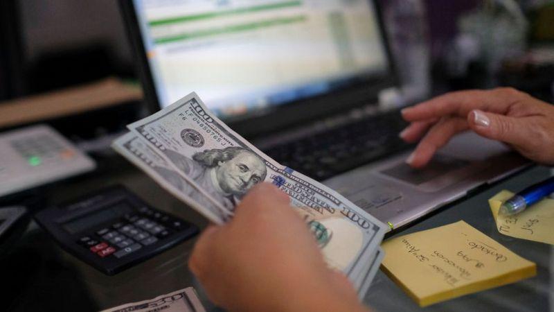 Mão de mulher digitando em laptop e segurando notas de dólar na outra