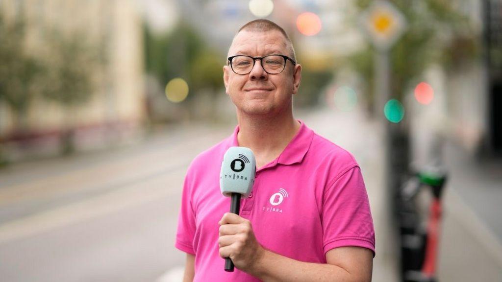 Fotografia colorida mostra homem branco de cabelo raspado e óculos, camisa rosa, segurando um microfone e sorrindo para a câmera