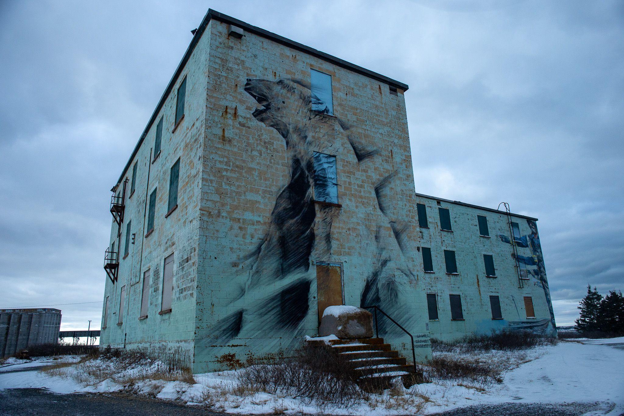 A imagem mostra um prédio antigo com um mural retratando um urso polar, em um ambiente frio e parcialmente coberto de neve.