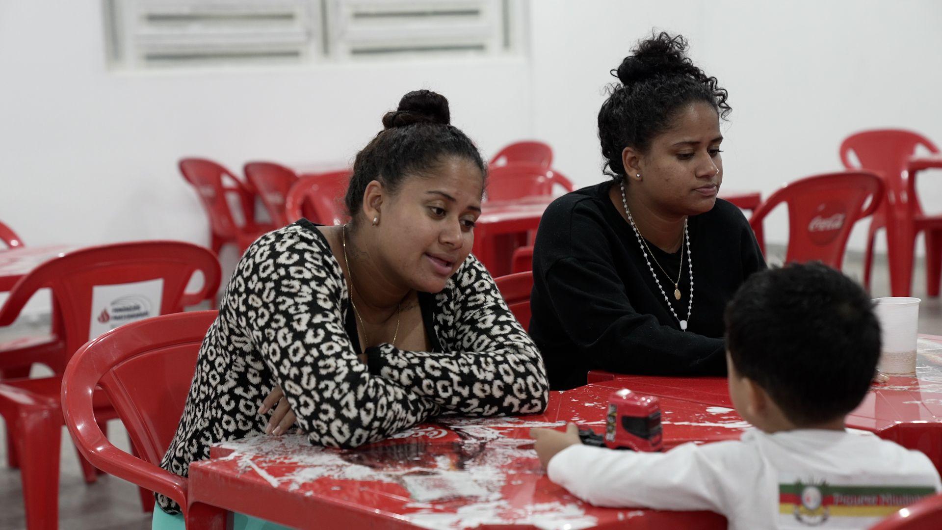 Duas mulheres sentadas diante de mesa e de criança de costas