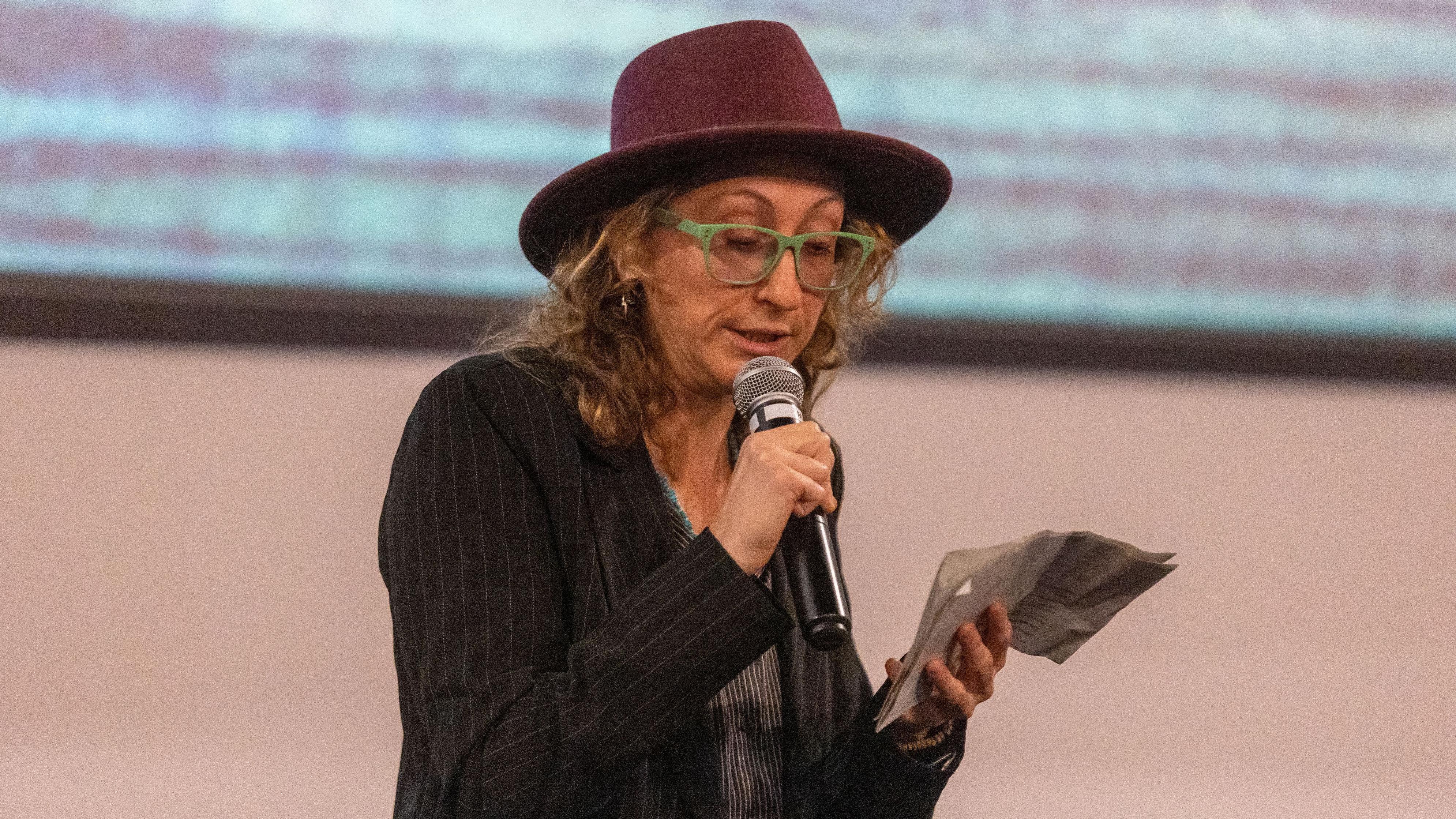 Halyna Hutchins' friend Rachel Manson reads from a piece of paper. She's wearing a red hat and speaks into the microphone.