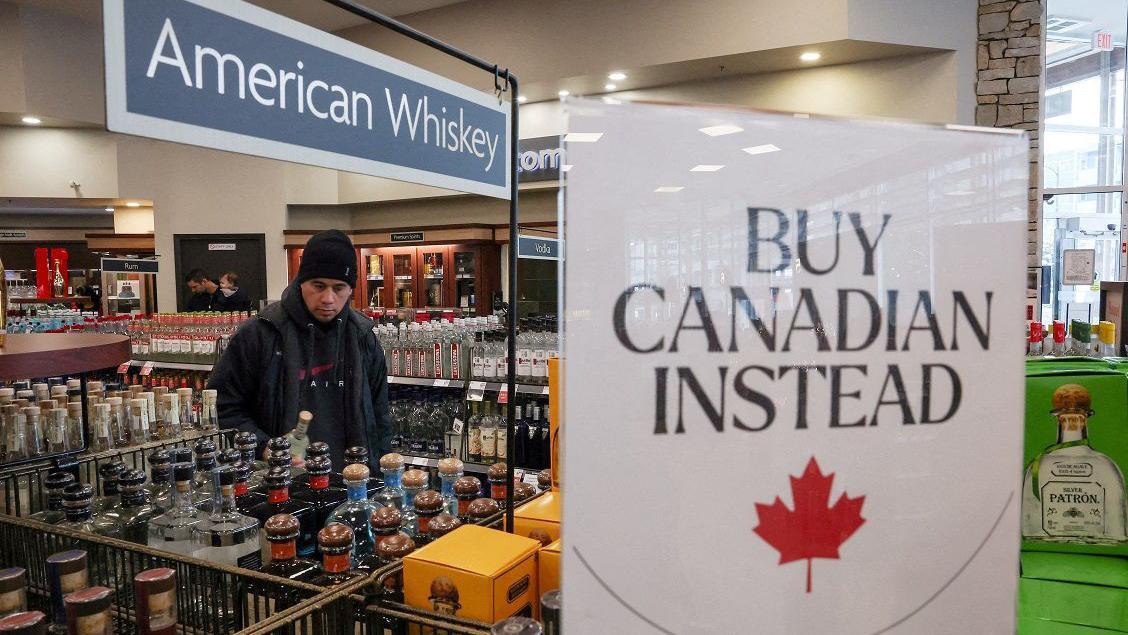 Um homem em uma loja de bebidas, onde há uma placa pedindo para comprar produtos canadenses. 