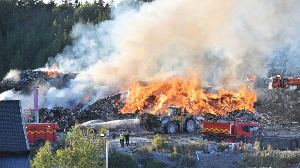 Carros de bombeiros são vistos ao lado de uma montanha de lixo em chamas